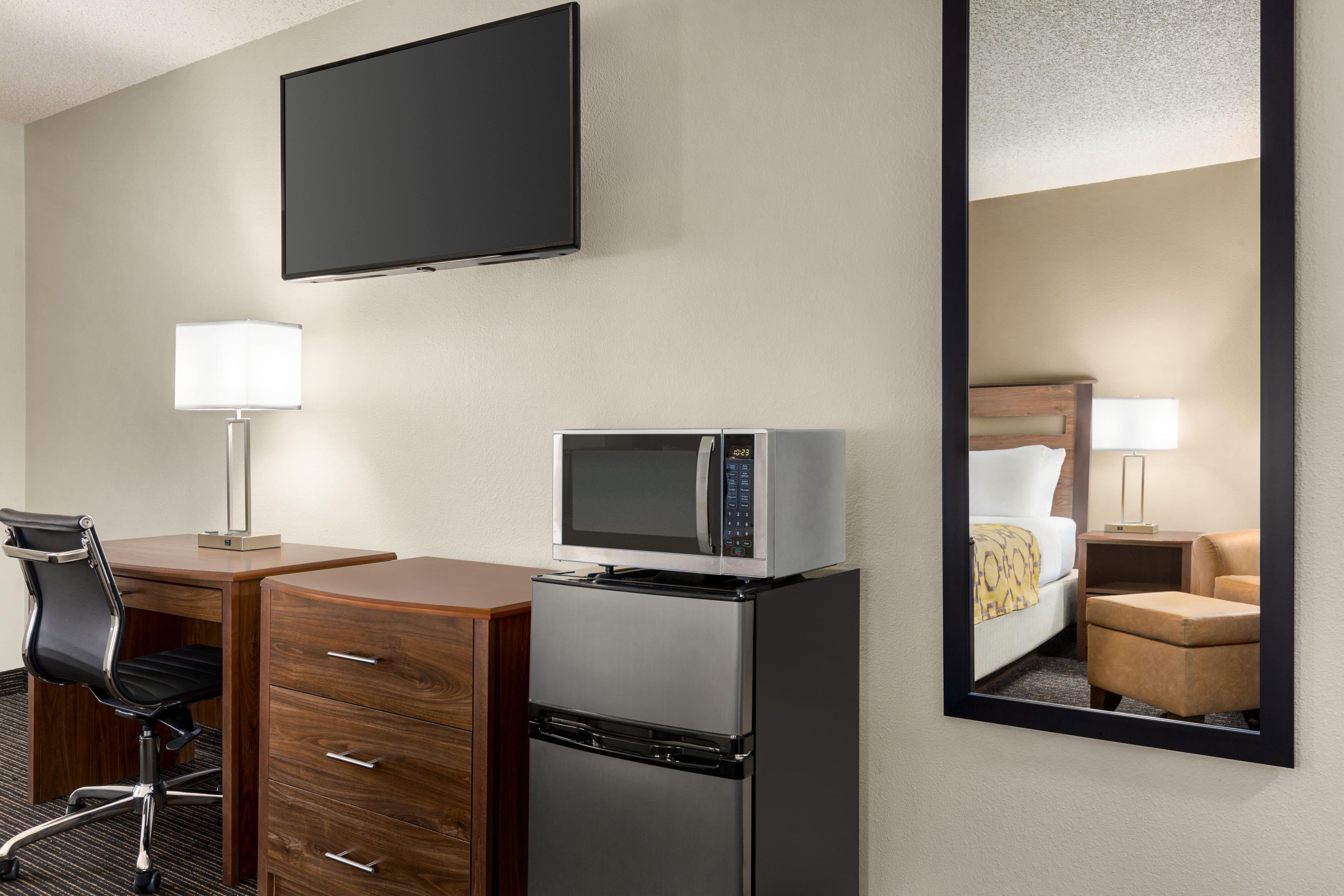 A hotel room with a flat-screen TV, a microwave, a refrigerator, a desk with a chair, a bedside lamp, and a large mirror.