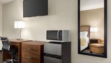 A hotel room with a flat-screen TV, a microwave, a refrigerator, a desk with a chair, a bedside lamp, and a large mirror.