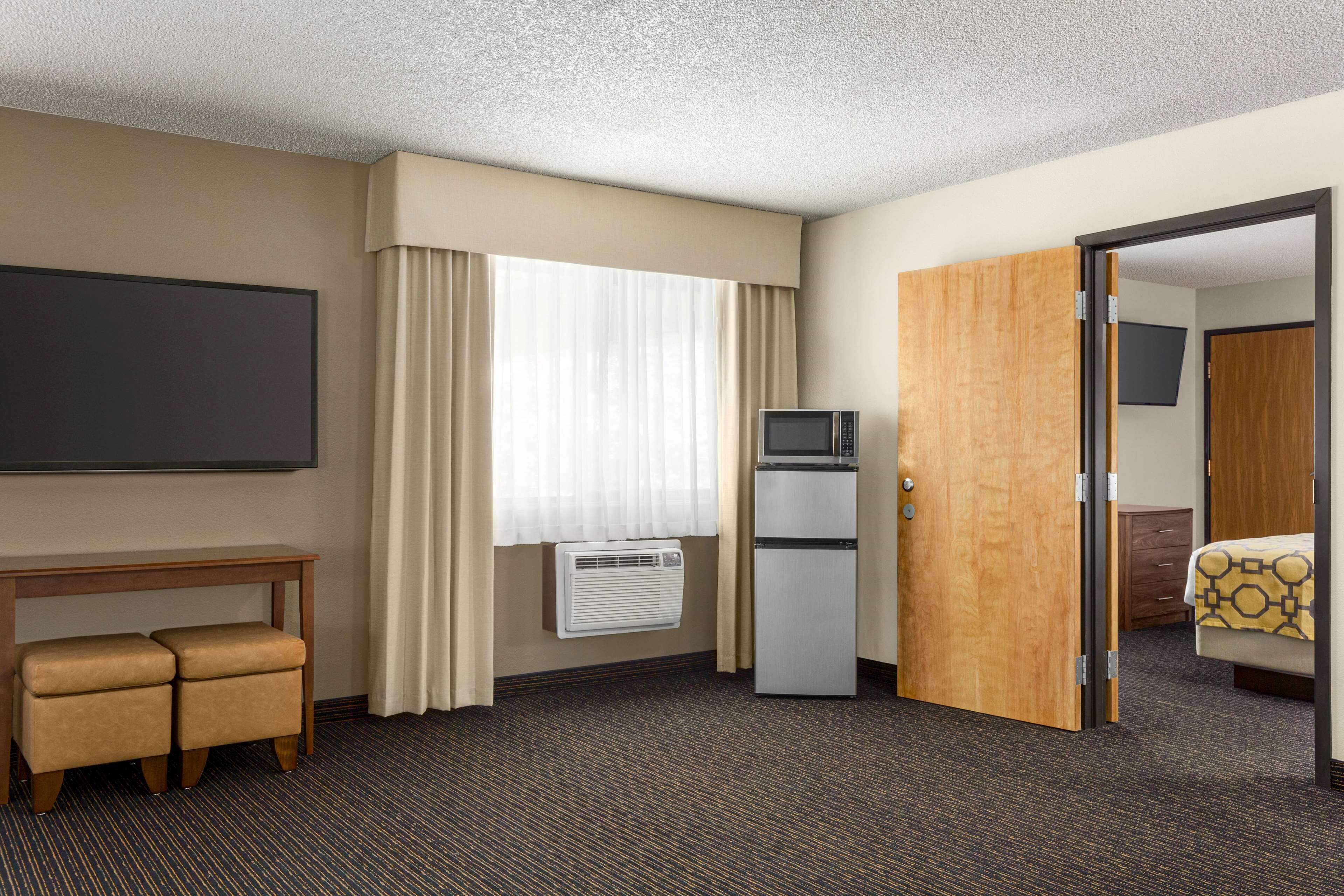 A hotel room with a bed, a desk with a chair, a television, a refrigerator, and a door leading to another room.