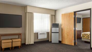 A hotel room with a bed, a desk with a chair, a television, a refrigerator, and a door leading to another room.