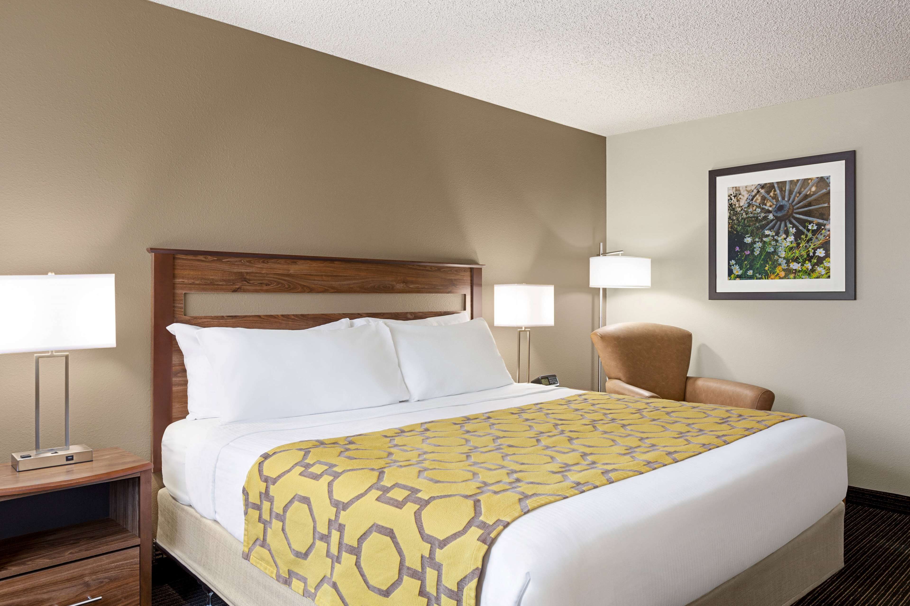 Habitación de hotel con cama, dos lámparas de noche, una silla, mesita de noche y un cuadro en la pared.