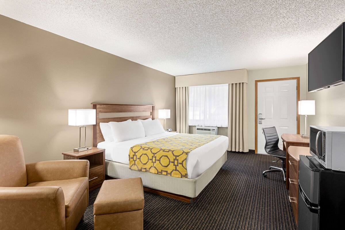A hotel room with a bed, two bedside tables with lamps, a desk with a TV, a chair, and a refrigerator.