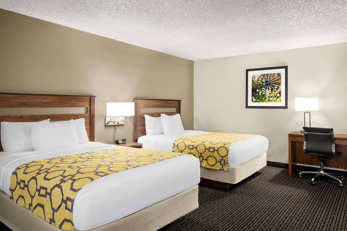 A hotel room with two beds, a desk, a chair, and a nightstand with a lamp.