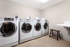 A laundry room with four front-loading washing machines, a small wooden stool, and a white sink. - Baymont by Wyndham Whitefish (Whitefish)