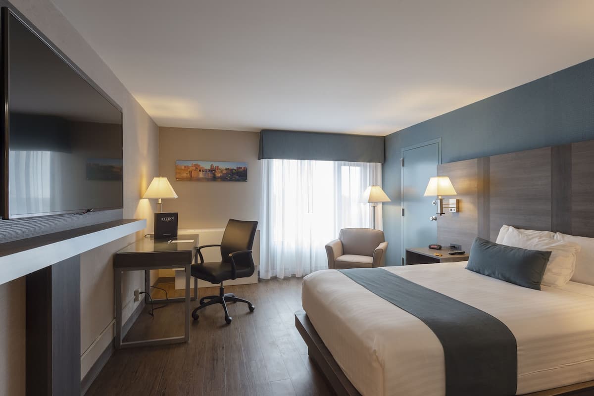 A hotel room with a large bed, a desk with a chair, two lamps, a TV, and a painting on the wall.