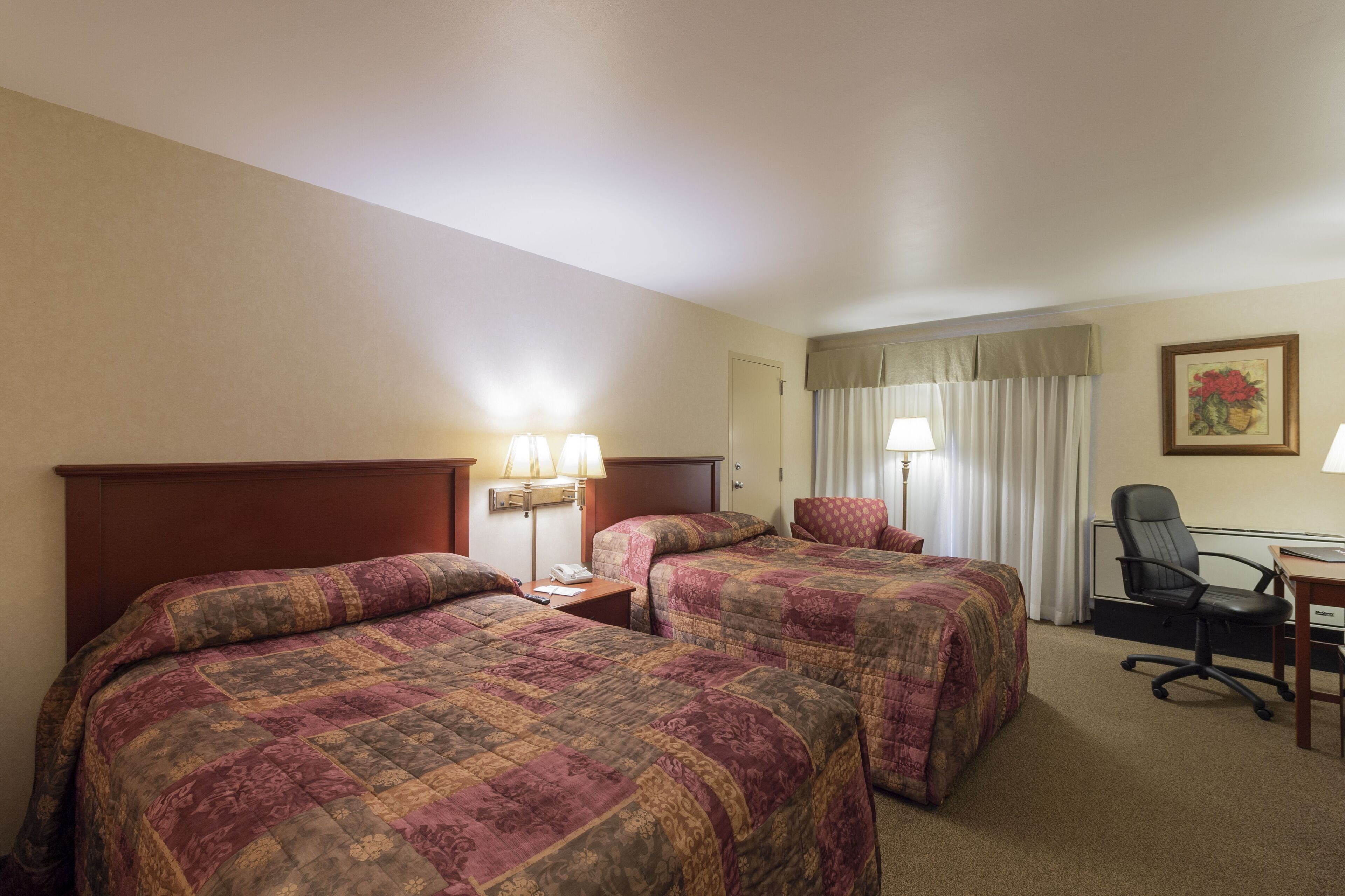 A hotel room with two beds, a desk, a chair, a lamp, and a painting on the wall.