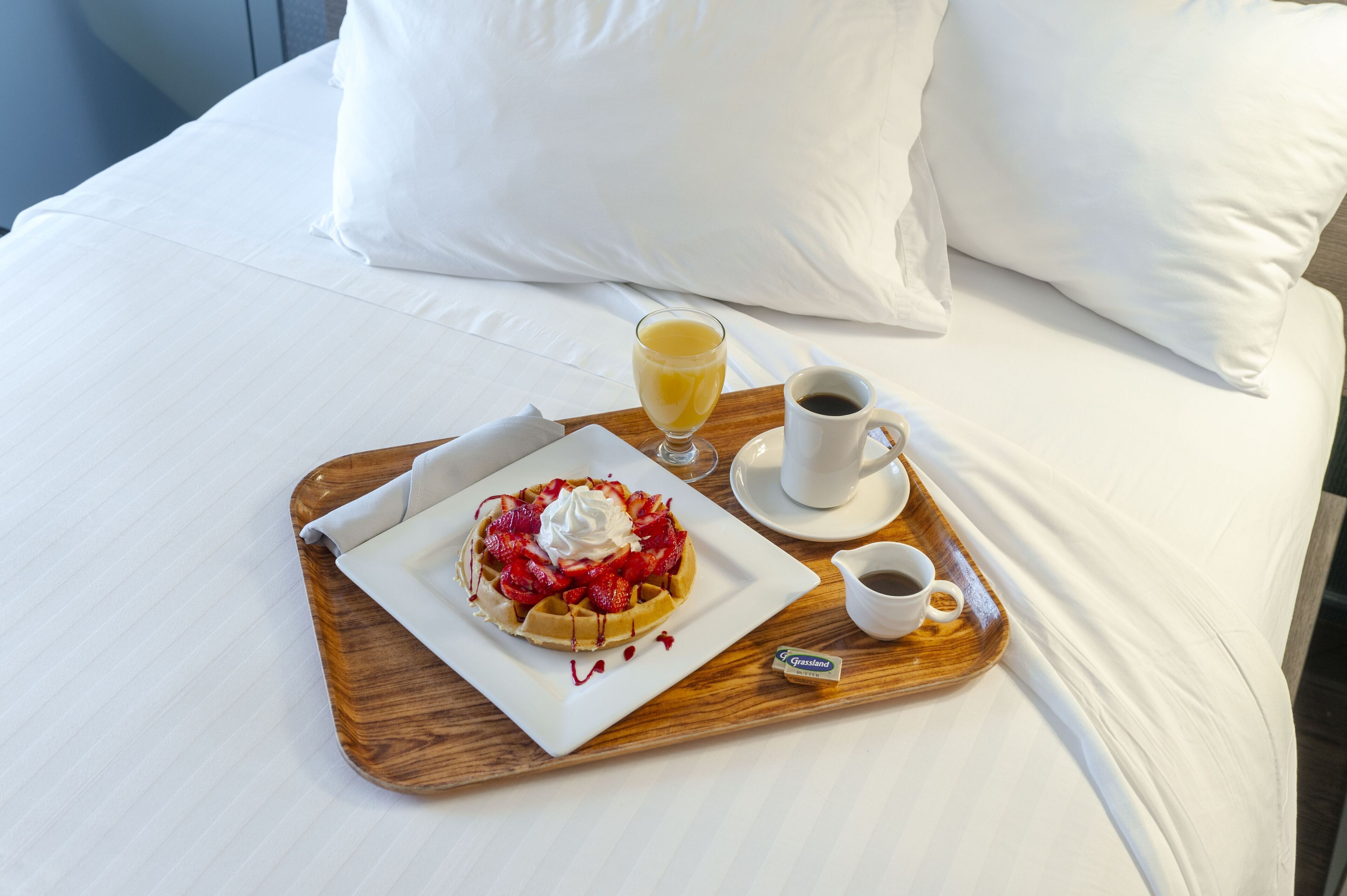 Bandeja de desayuno con waffles, crema batida, fresas y jarabe sobre una cama con ropa blanca y almohadas.