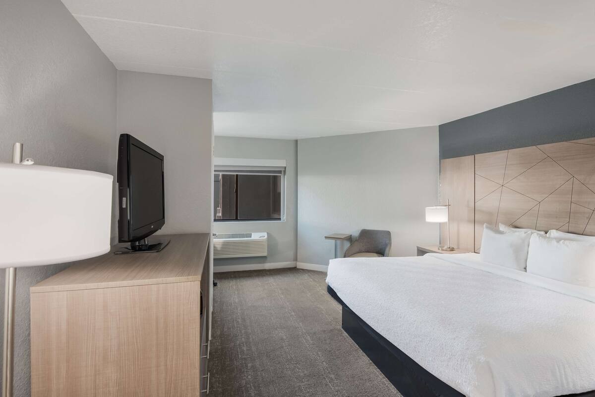 A hotel room with a bed, a TV on a wooden desk, a bedside table with a lamp, and a window with a view.
