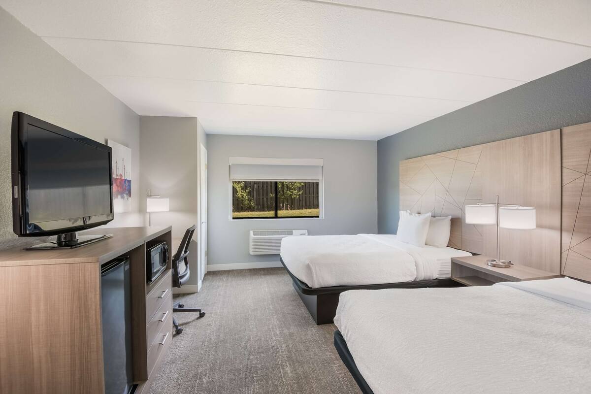 A hotel room with two beds, a TV, a desk with a microwave, and a window with a view of trees.