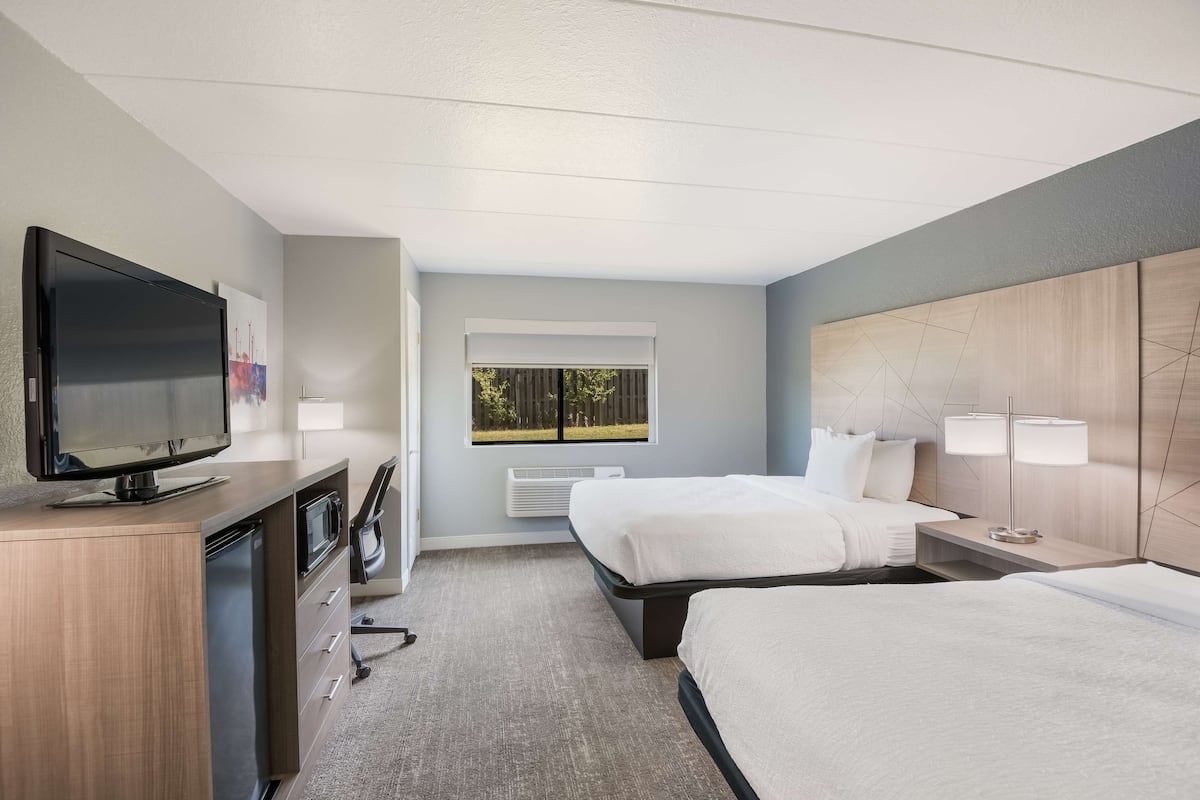 Ein Hotelzimmer mit zwei Betten, einem Fernseher, einem Schreibtisch mit Mikrowelle und einem Fenster mit Blick auf Bäume.
