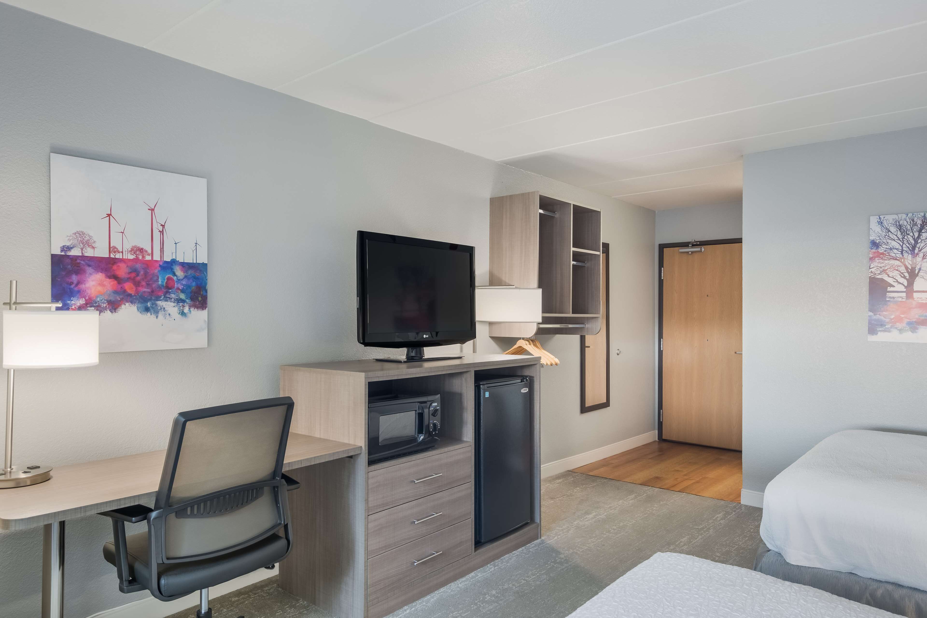 A modern hotel room with a desk, a television, a mini-fridge, and a bed.