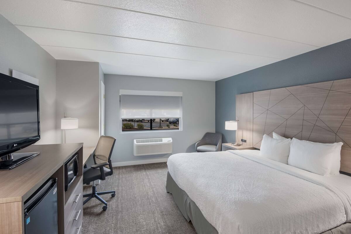 A hotel room with a bed, a desk with a TV, a chair, a window with a view, and a wall-mounted air conditioner.