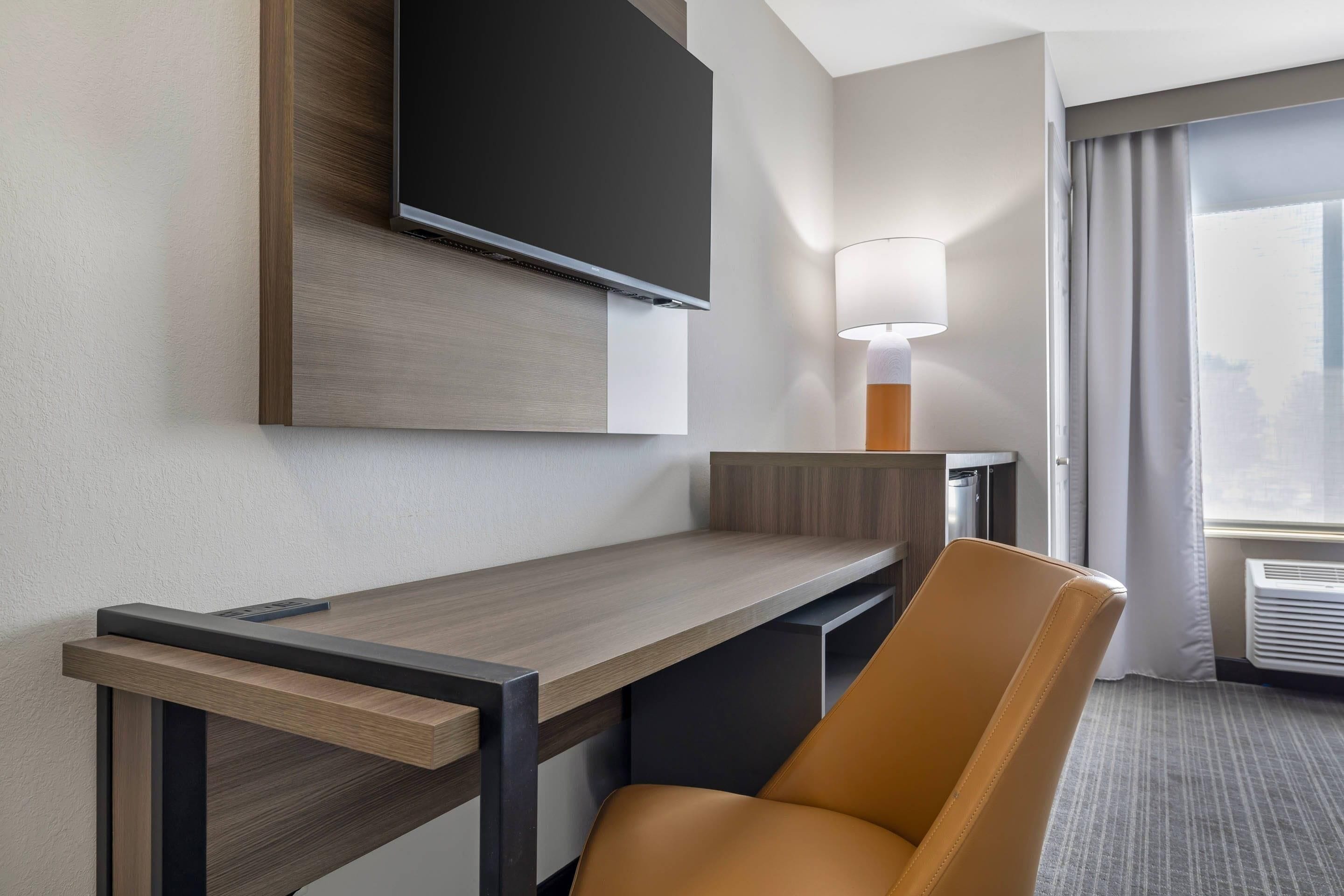 A hotel room with a flat-screen TV mounted on the wall, a desk with a chair, a lamp, and a view of the city through the window.