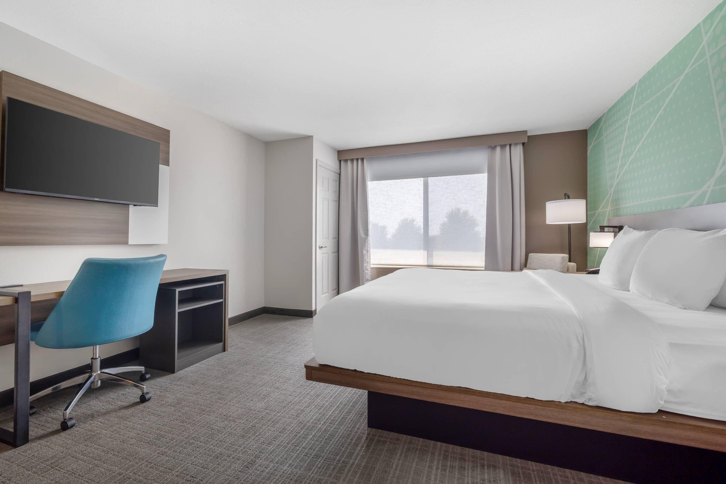 Una habitación de hotel con cama, escritorio, silla, televisor y ventana con cortinas.