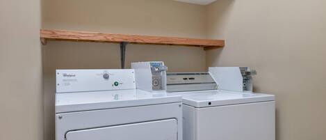 A laundry area with two Whirlpool washers and a dryer, a wooden shelf above, and a white ceiling.