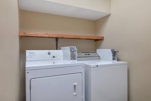 A laundry area with two Whirlpool washers and a dryer, a wooden shelf above, and a white ceiling.