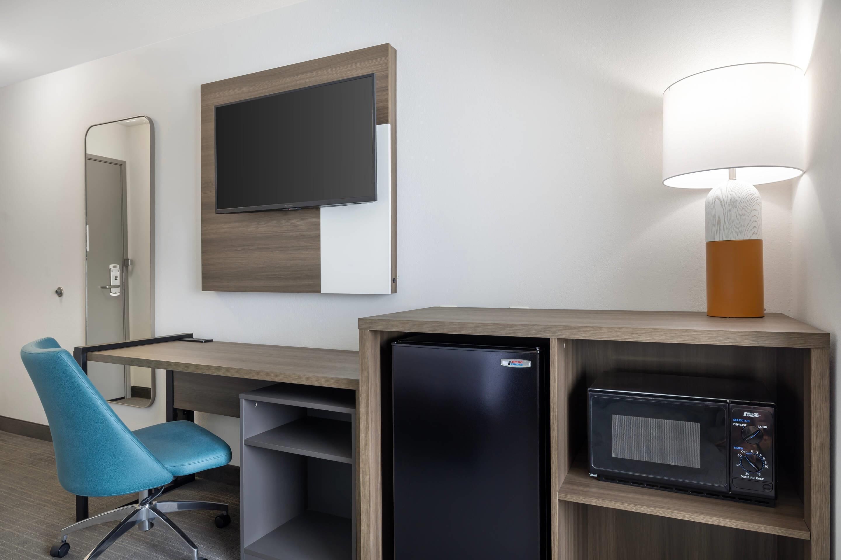 A hotel room with a desk, a television mounted on the wall, a microwave, a lamp, a chair, and a refrigerator.
