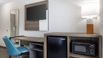 A hotel room with a desk, a television mounted on the wall, a microwave, a lamp, a chair, and a refrigerator.