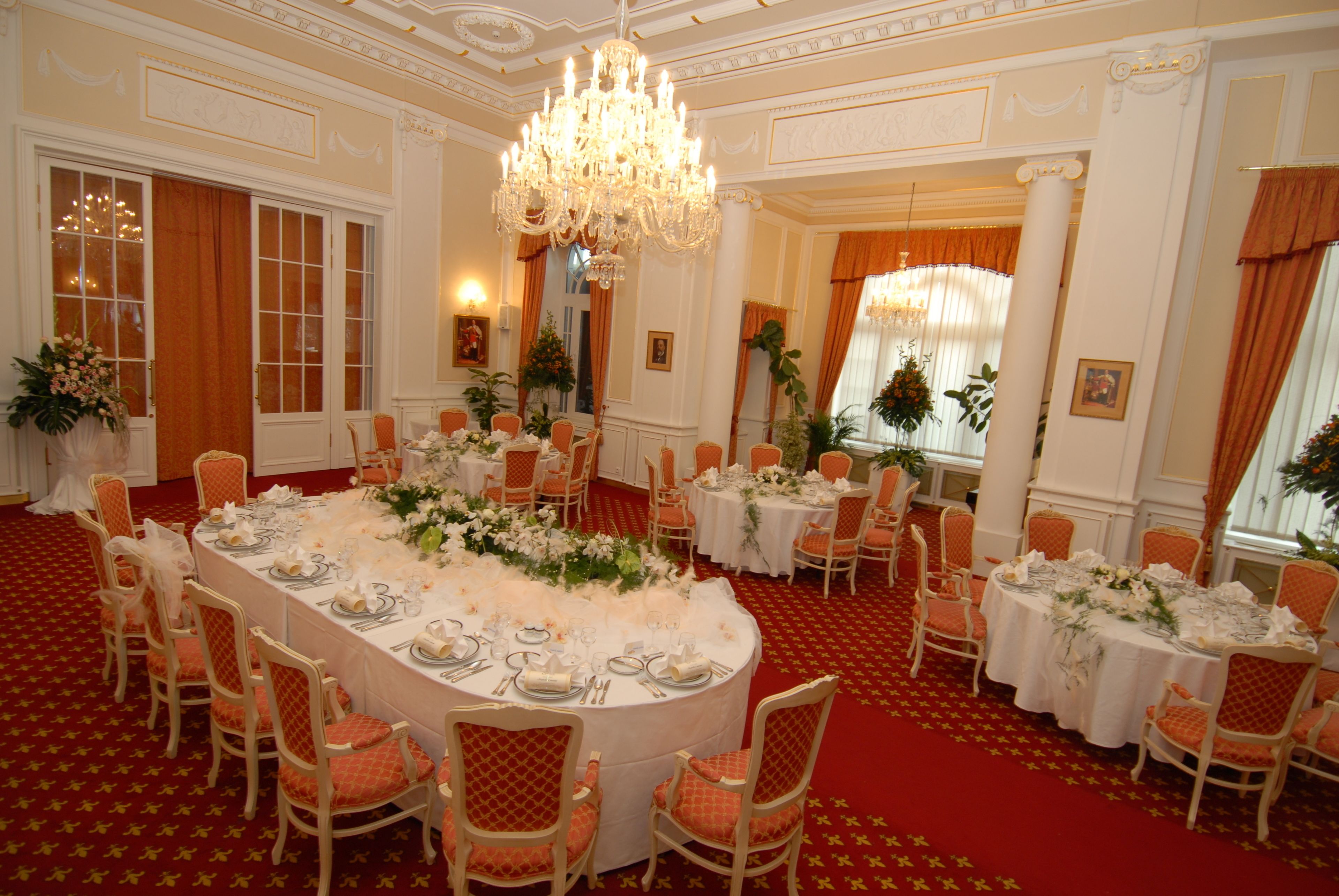 A formal dining room with round tables, white tablecloths, and orange chairs.
