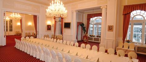 A banquet hall with a long table set for a formal event, chandeliers, and classical architecture.