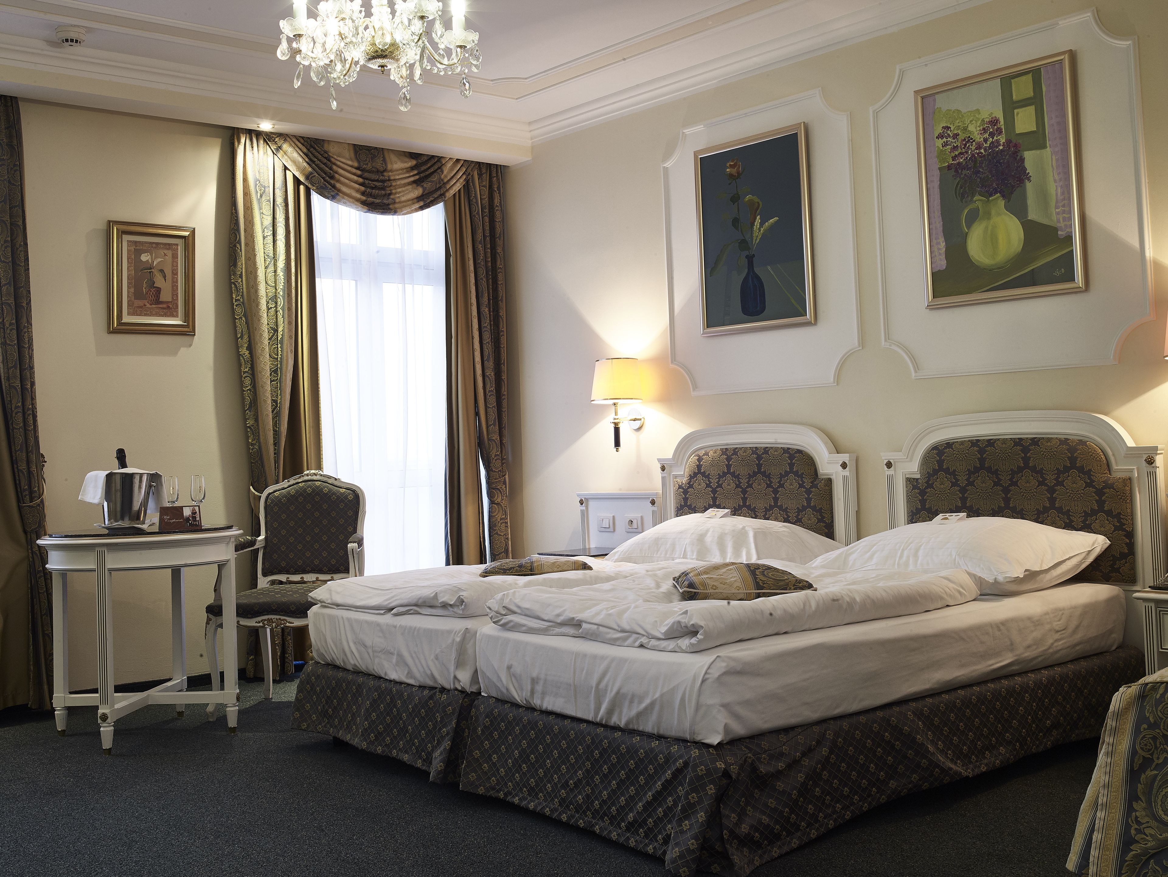 A hotel room with a large bed, a chair, a small table with a pitcher and glasses, and two paintings on the wall.