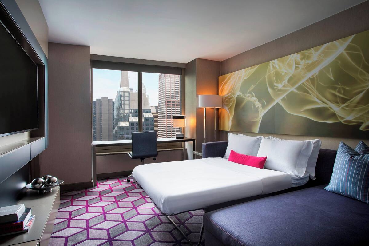 Habitación de hotel con cama, escritorio, silla, televisor y ventana grande con vista a la ciudad.