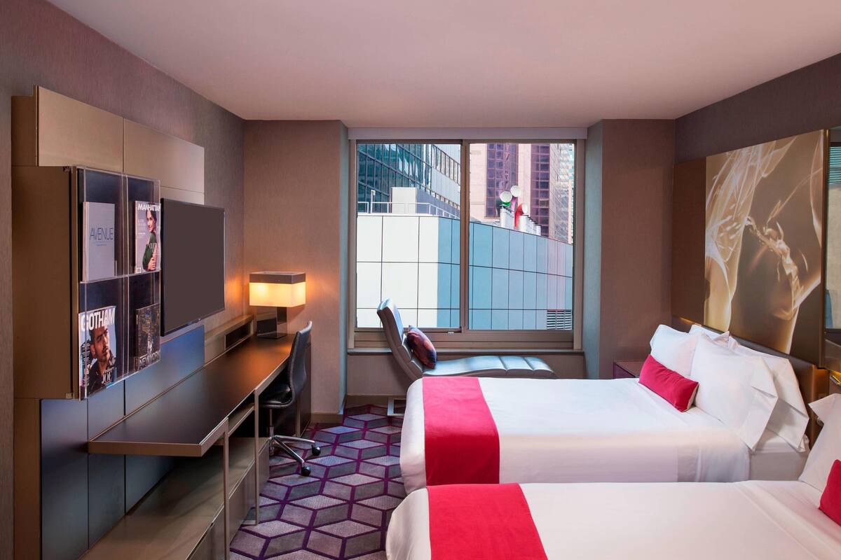 Habitación de hotel con dos camas, un escritorio, una silla, un ventanal grande y un televisor montado en la pared.