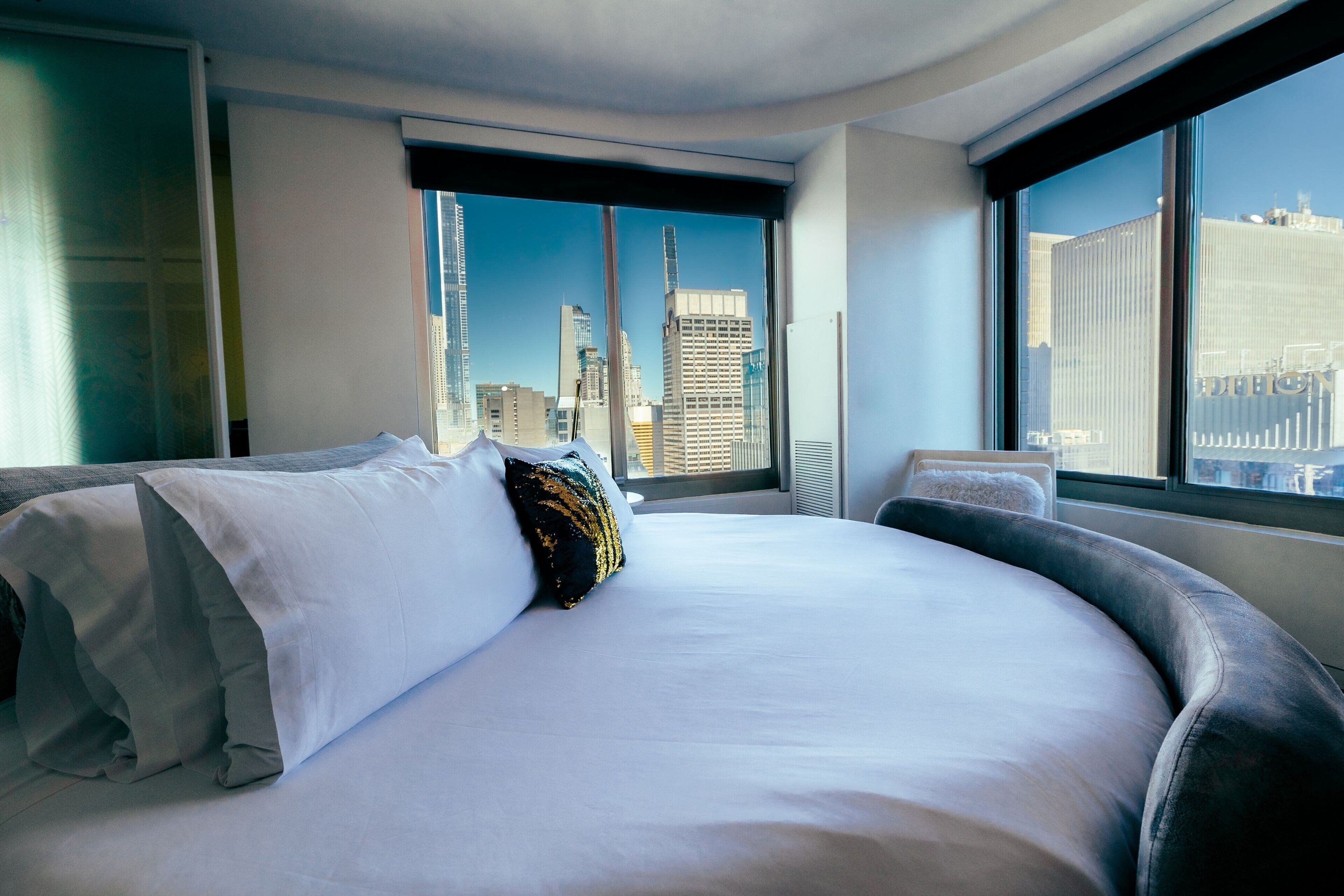 A modern hotel room with a large bed, a view of the city skyline, and a window with a black frame.