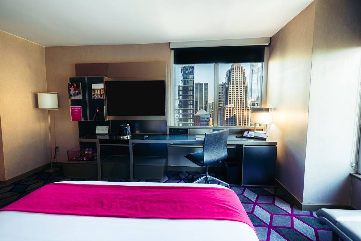 Habitación de hotel con cama, escritorio, silla, televisor y vista a la ciudad.