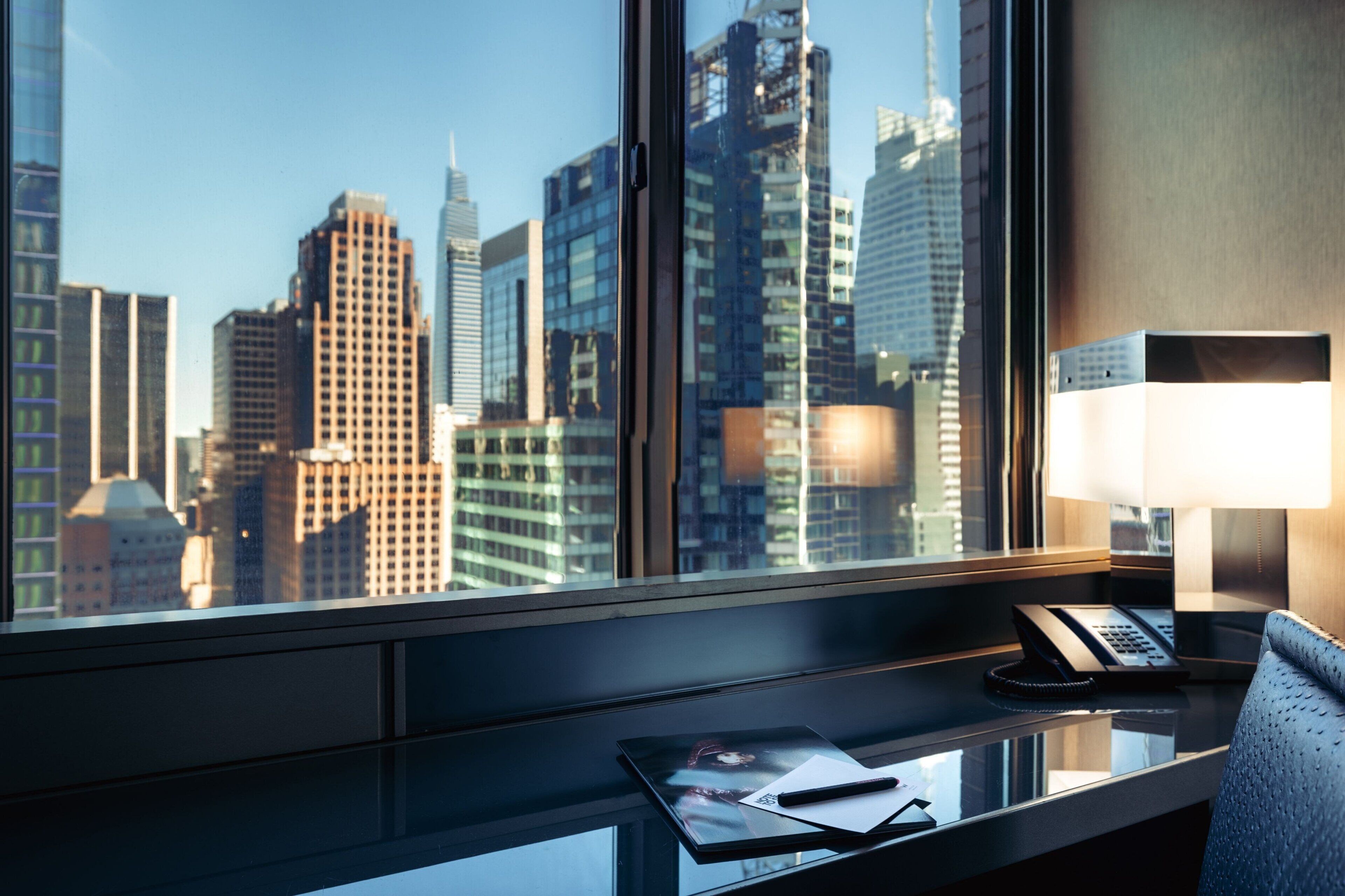 Ein Hotelzimmer mit Schreibtisch, Lampe, Telefon und Fenster mit Blick auf die Stadt.