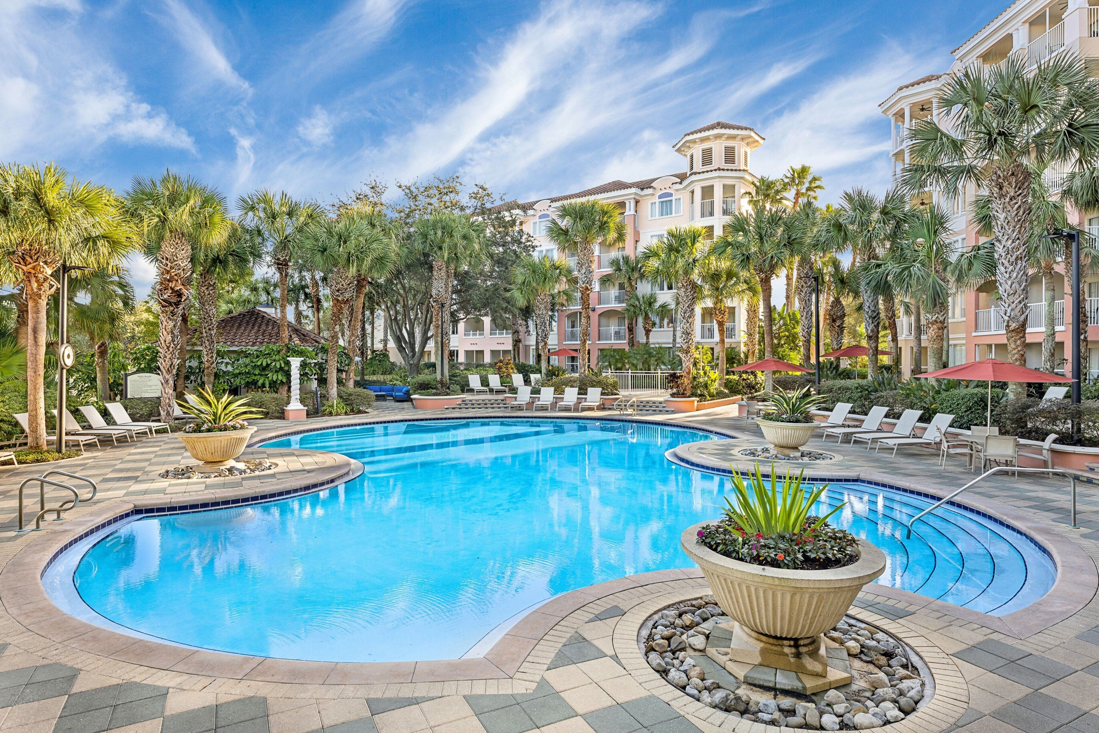 A pool area with a curved pool, lounge chairs, palm trees, and a building with balconies in the background.
