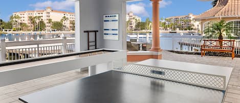 A ping pong table with two paddles, a ladder, and a sign for "The Boathouse" in a resort setting with a view of a pool and buildings.