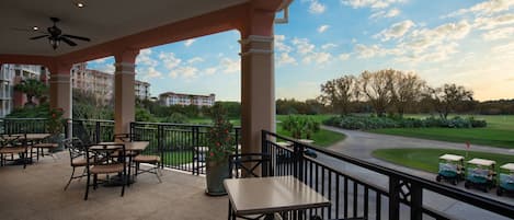 A covered outdoor area with tables and chairs, a view of a golf course, and buildings in the background.