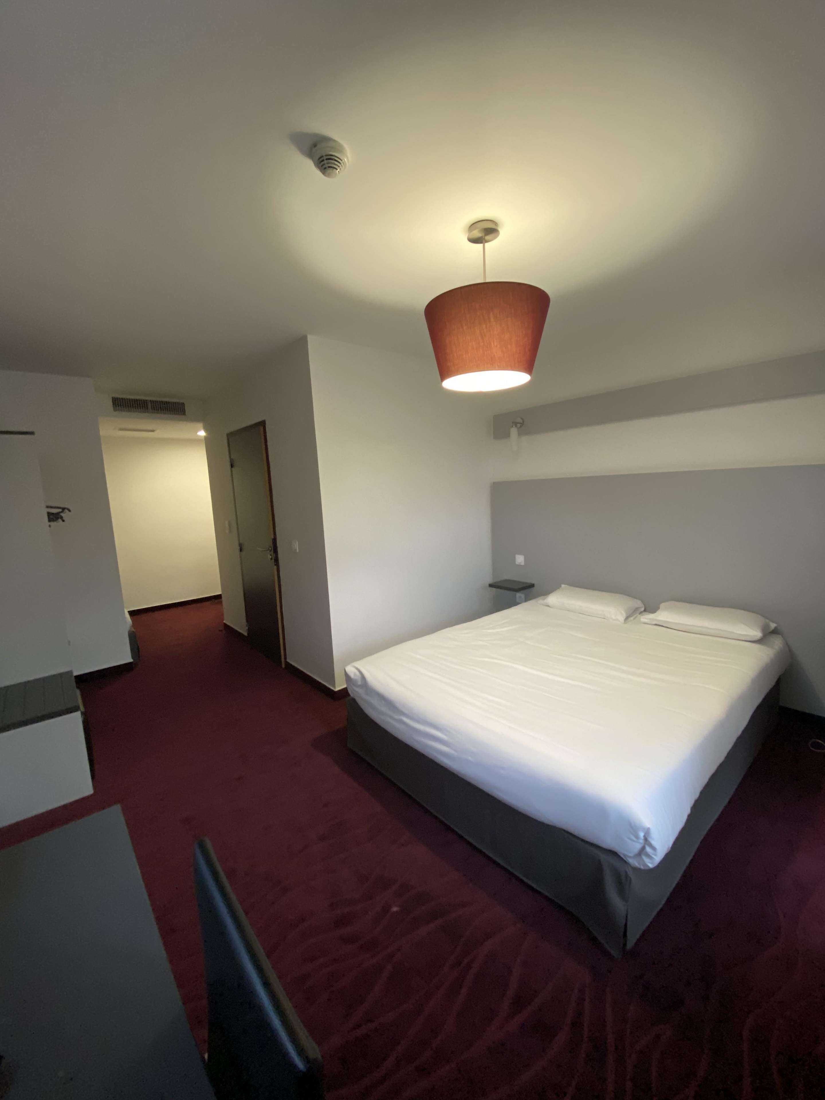 A hotel room with a bed, a desk, a chair, a red carpet, and a ceiling light.