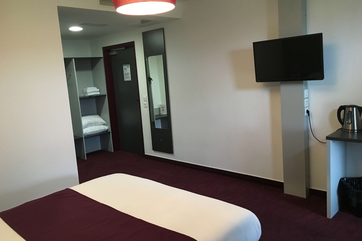 Une chambre d’hôtel comprenant un lit, un téléviseur fixé au mur, une petite table avec une bouilloire et une porte donnant sur un placard.