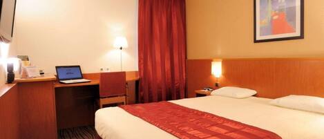 A hotel room with a bed, a desk with a laptop, a chair, a nightstand, and a painting on the wall.