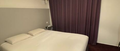 Una habitación de hotel con una cama grande, una mesita de noche, una lámpara y cortinas oscuras.