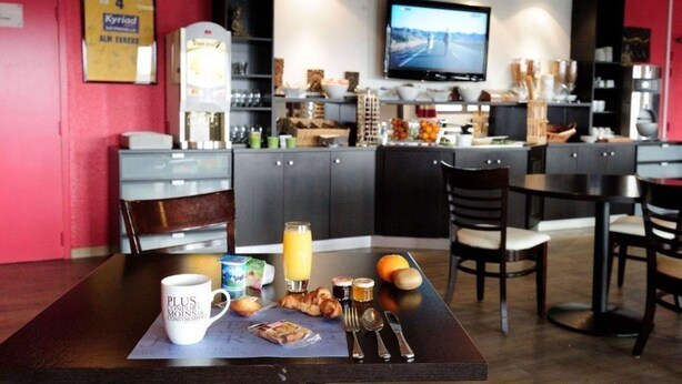 Een ontbijtje met een koffiemok, croissants en sinaasappelsap op een tafel, met een televisie op de achtergrond en een rode muur.