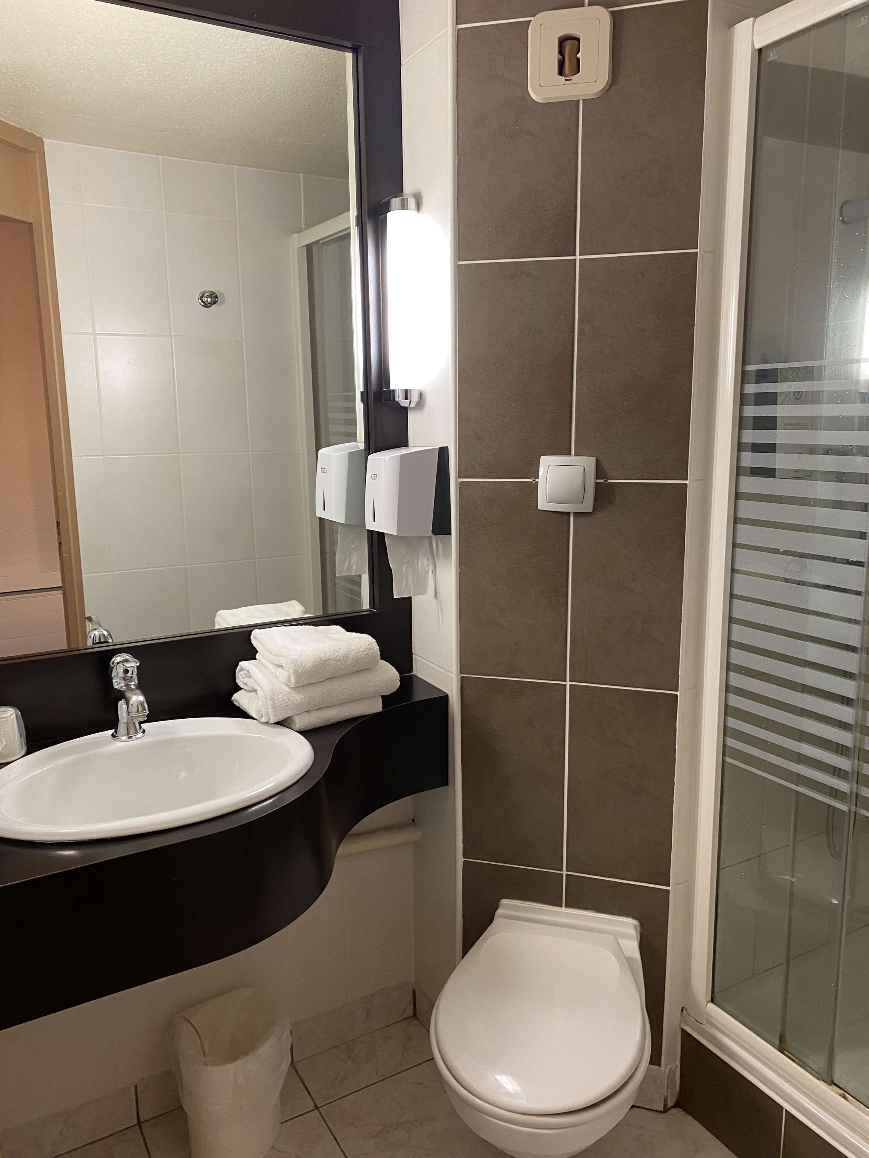 A bathroom with a sink, mirror, towel dispenser, toilet, and a glass shower enclosure.
