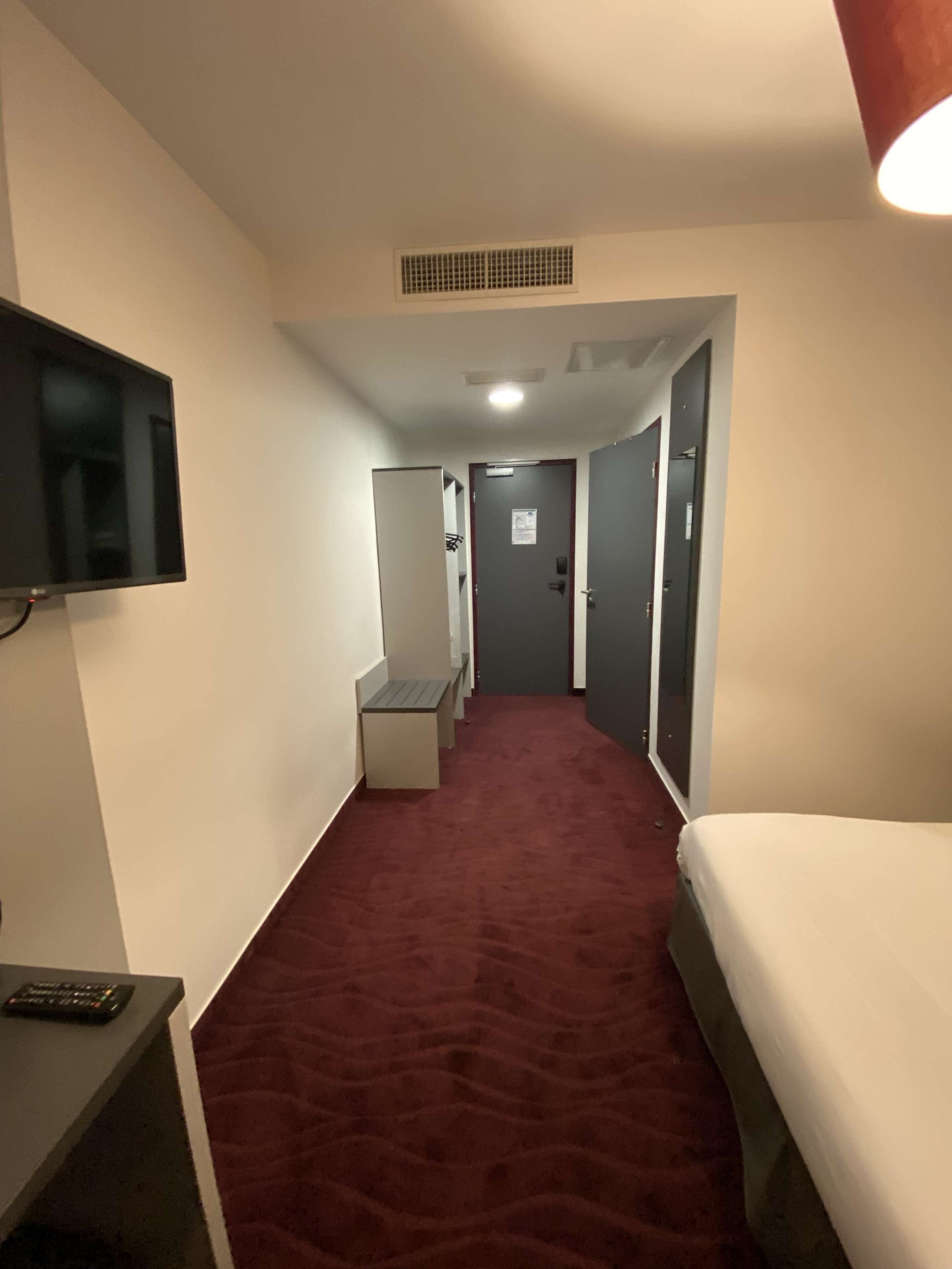 Habitación de hotel con alfombra roja, una cama, televisor montado en la pared y un escritorio con una silla.