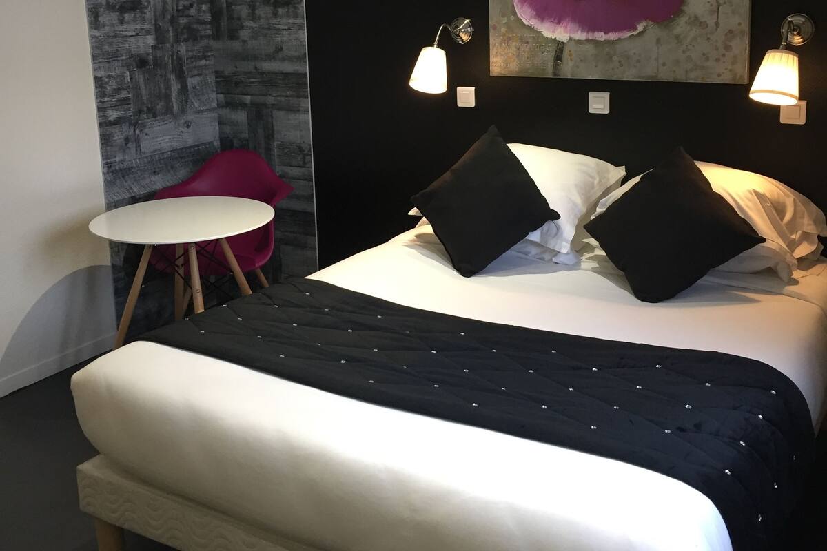 A modern hotel room with a large bed, a small round table, a red chair, and a large floral painting on the wall.