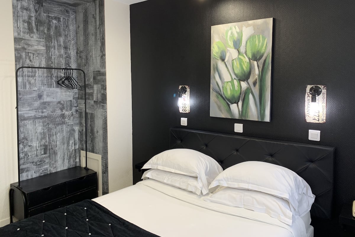 A bedroom with a black headboard, white bedding, a wall-mounted painting of green leaves, and a black wardrobe.
