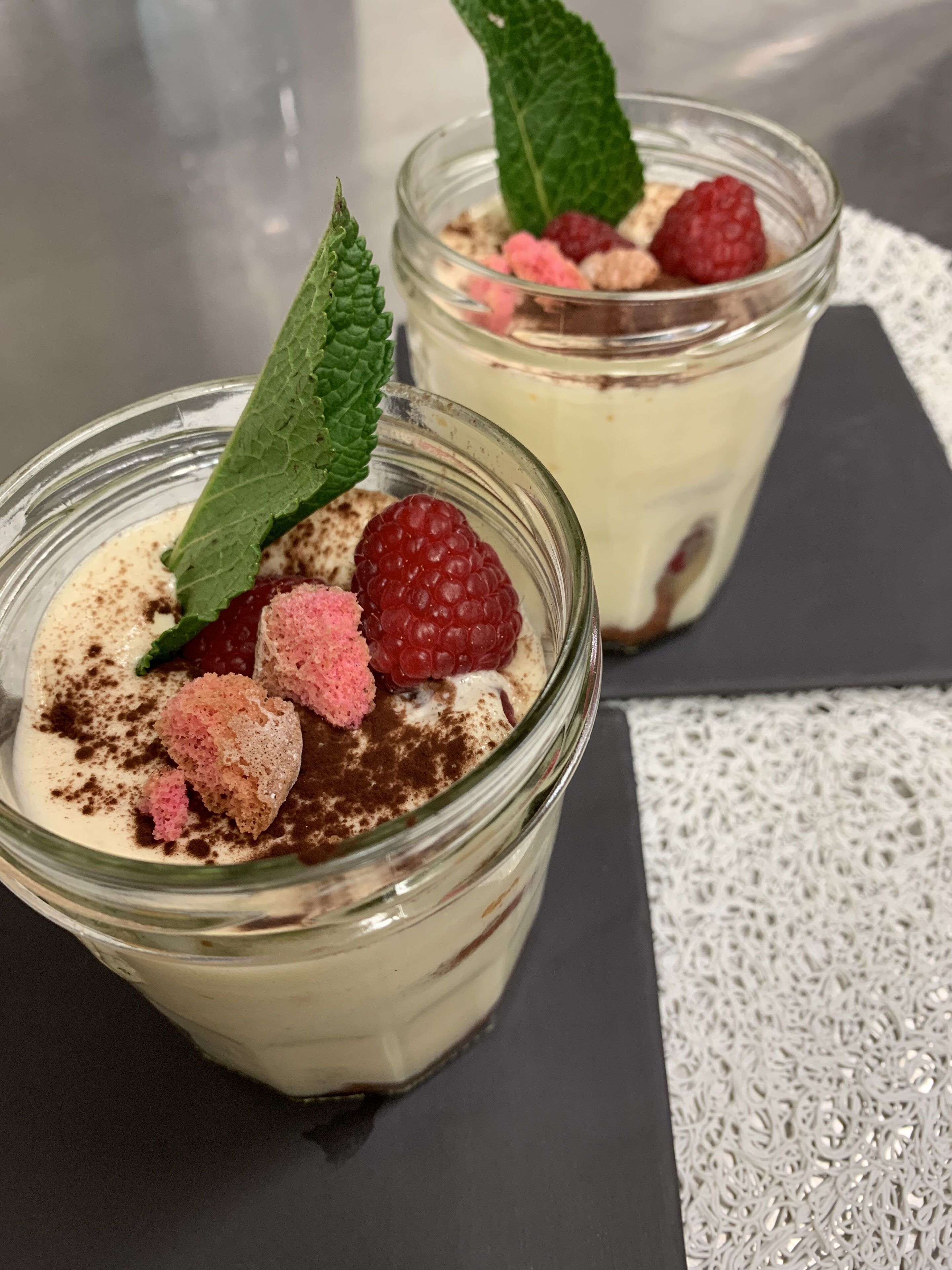 Deux coupes à dessert en verre, avec une base crémeuse, garnies de framboises, de sucre rose et d'une feuille de menthe.