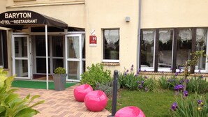 A hotel entrance with a glass door, a sign reading "BARYTON HOTEL RESTAURANT", and a small garden with potted plants and a bench.