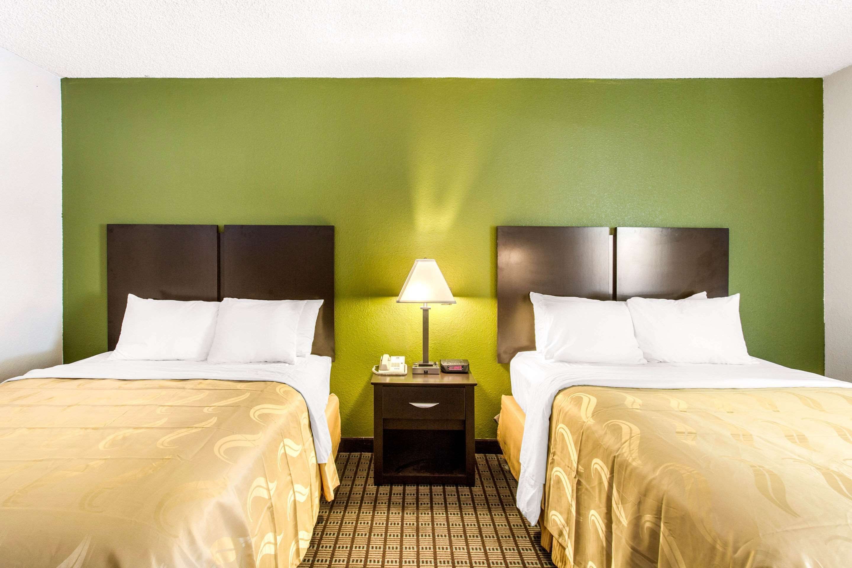 A hotel room with two beds, a nightstand with a lamp, and a green wall.