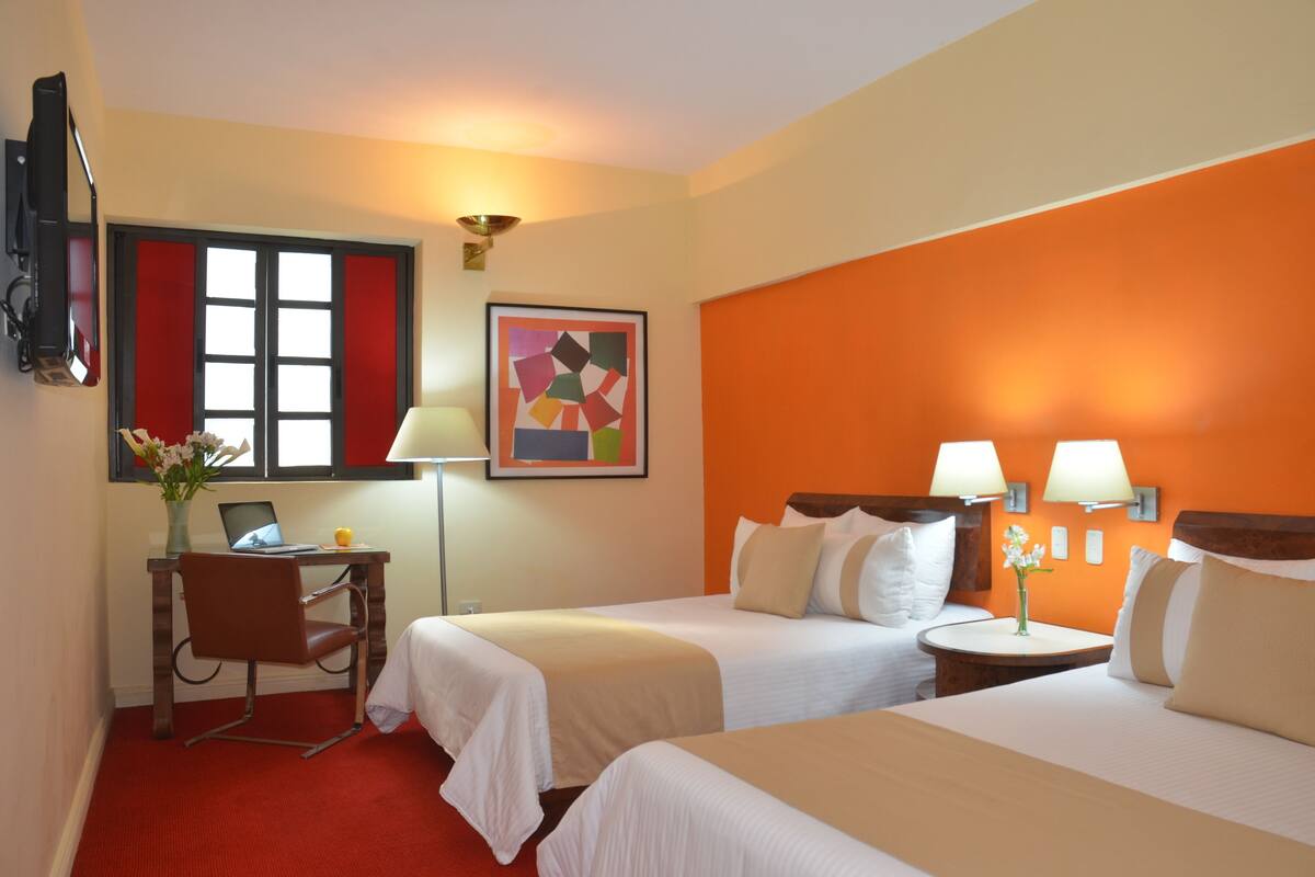 A hotel room with two beds, a desk with a laptop, a chair, a lamp, and a painting on the wall.