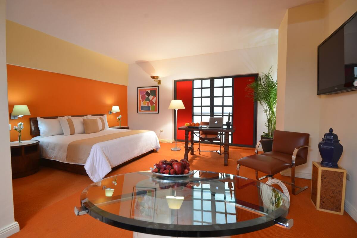A hotel room with a bed, a glass coffee table with fruit, a desk with a laptop, a chair, and a TV on the wall.