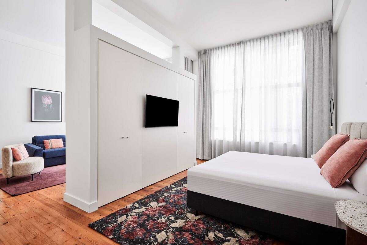 A modern bedroom with a large bed, a TV mounted on a white wall, a blue sofa, and a small armchair.