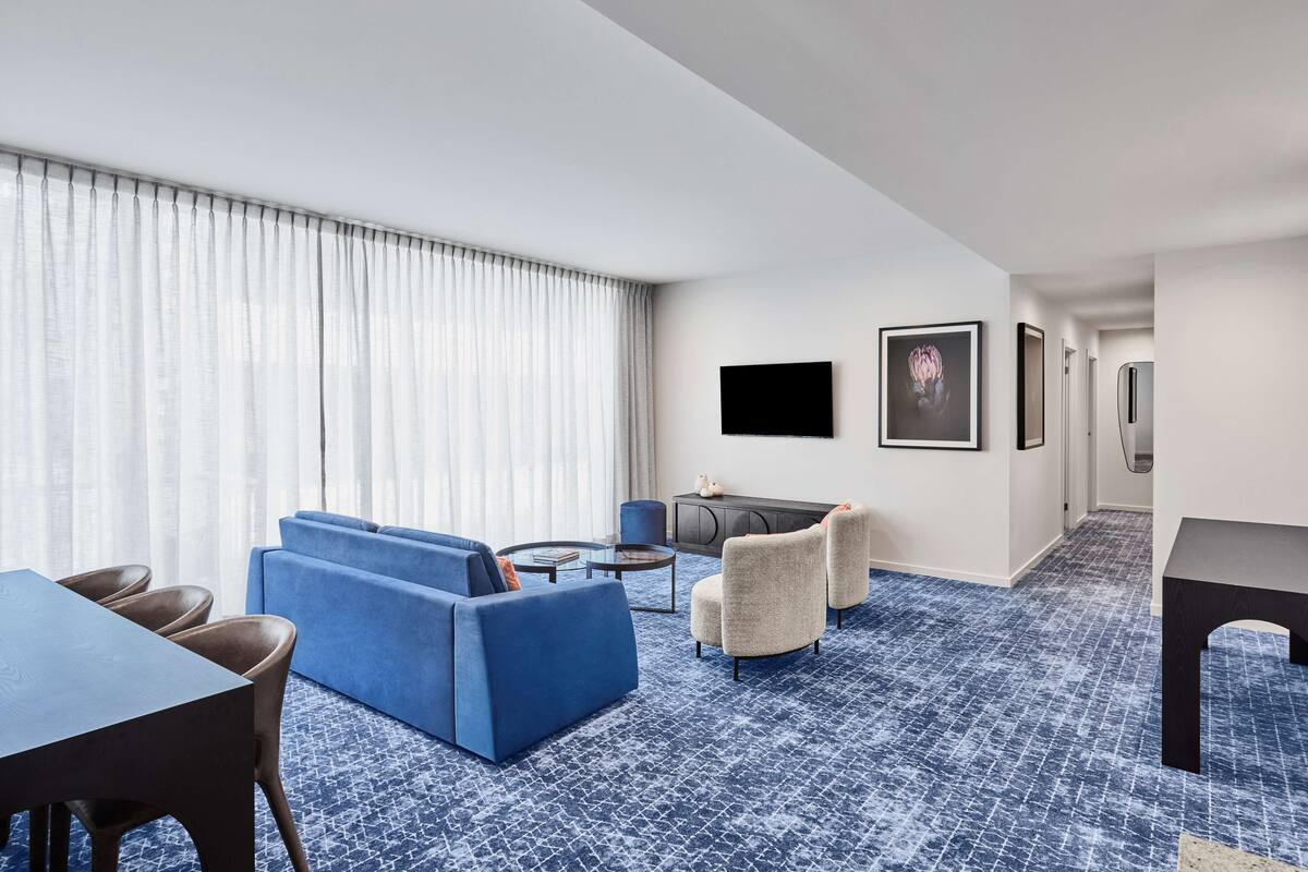 A modern hotel room with a blue patterned carpet, a blue sofa, a dining table with chairs, a small round coffee table, and a TV mounted on the wall.