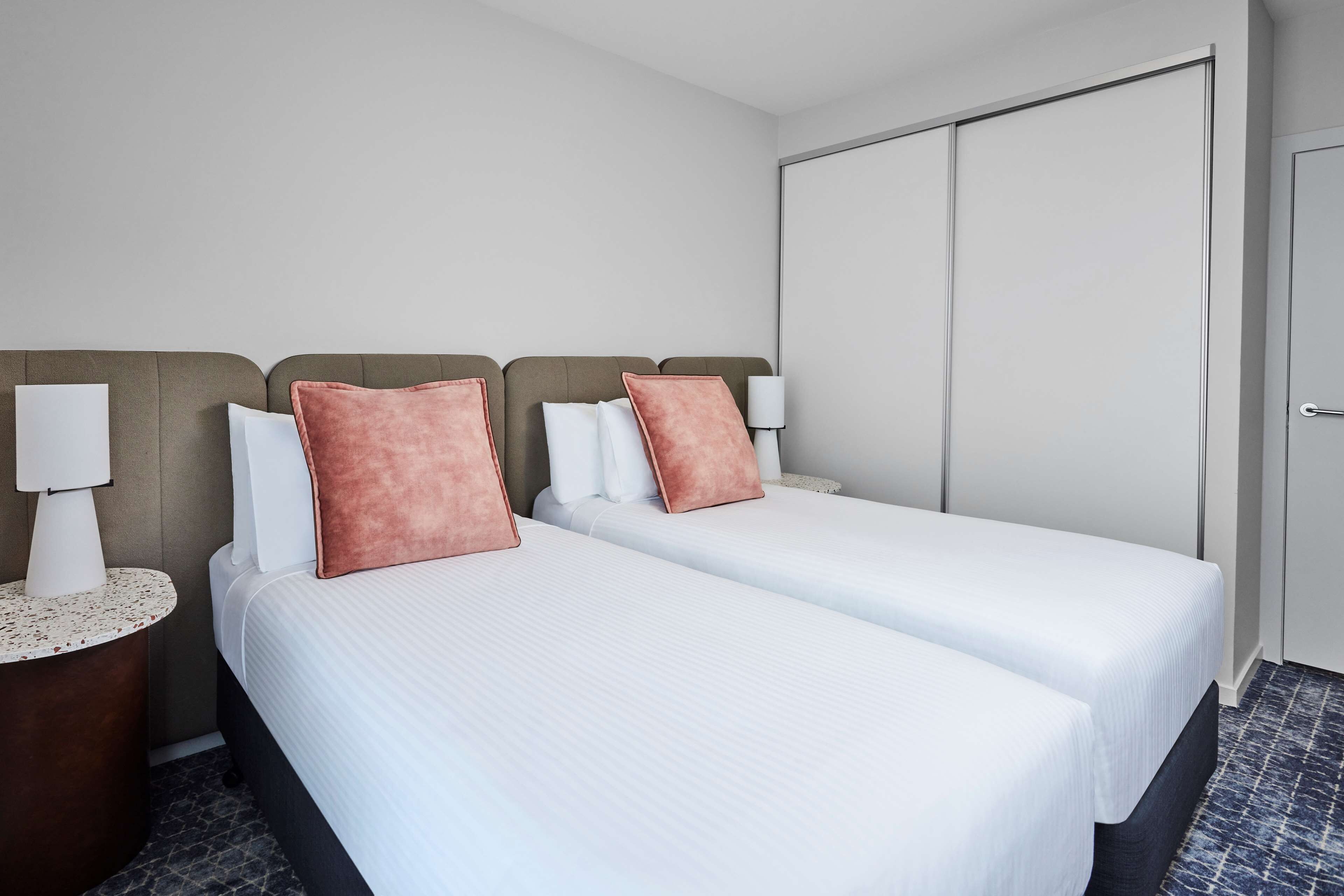 A hotel room with two beds, each with white bedding and a single rose gold pillow. There is a nightstand with a lamp on each side of the beds. The room has a large closet and a door to the right.